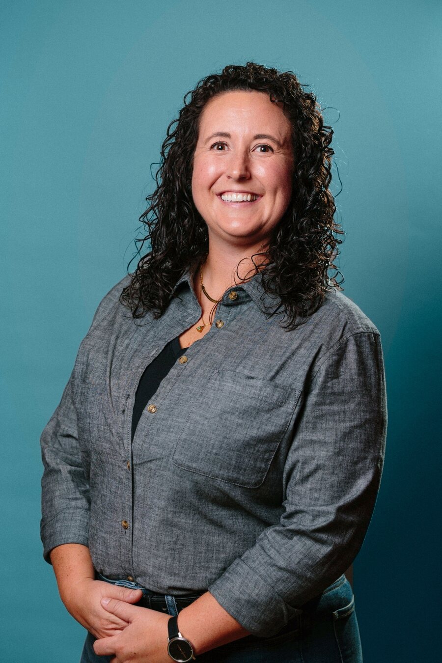 Portrait of Jillian van der Geest smiling, with long dark curly hair, wearing a grey chambray shirt against a teal background